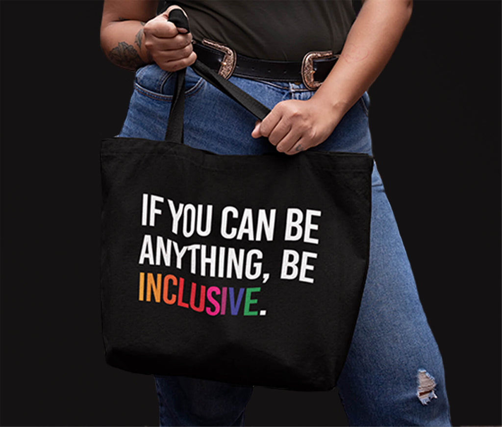 A close up photo of a person holding a black tote bag with the phrase, "If you can be anything, be inclusive." on it.