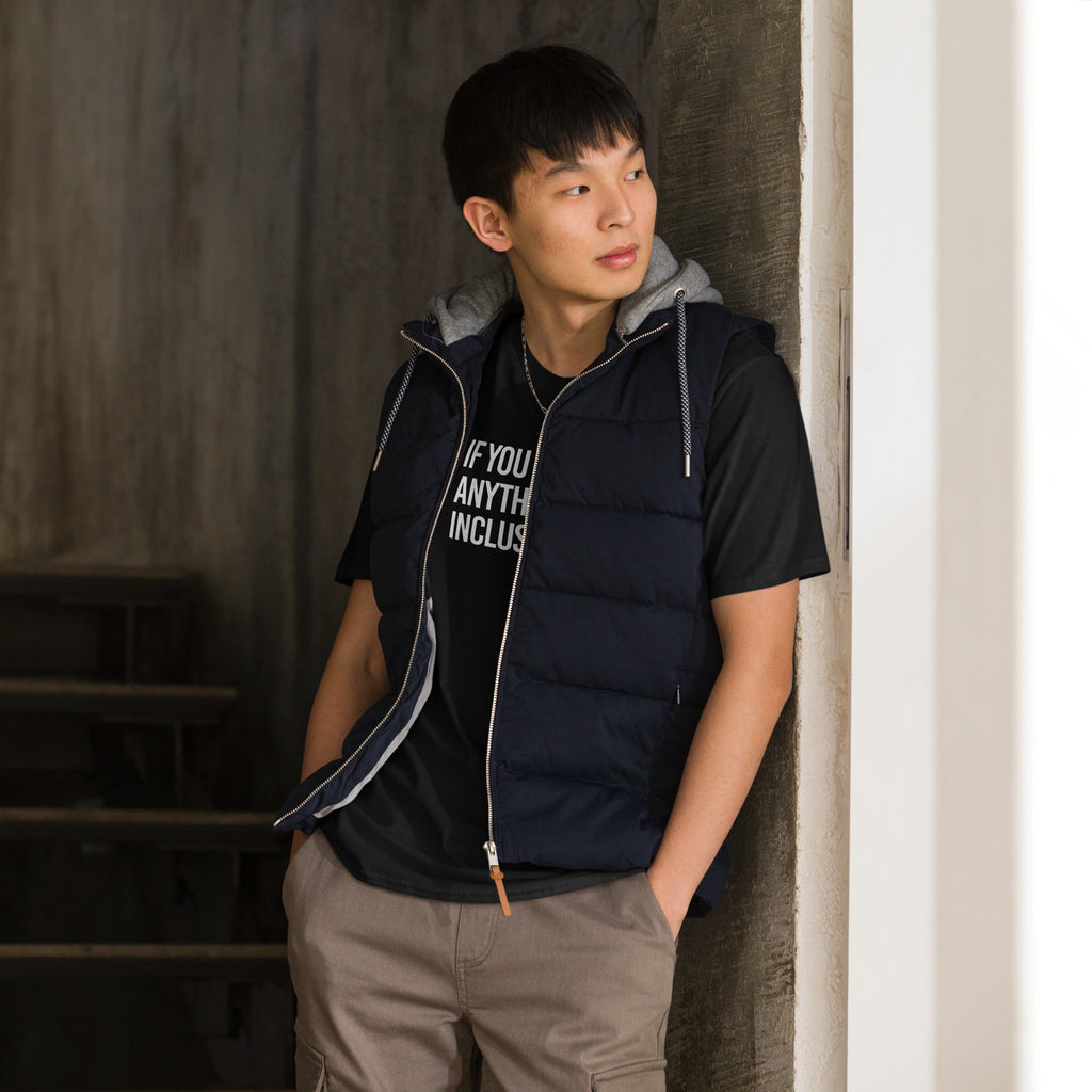 This is the photo of a black Be Inclusive Basic Tee. On the front of the t-shirt, the phrase, 'If you can be anything, be inclusive,' is printed in white capital letters. A man is wearing a puffy jacket over the tee.