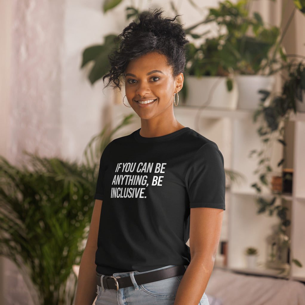 This is the photo of a black Be Inclusive Basic Tee. On the front of the t-shirt, the phrase, 'If you can be anything, be inclusive,' is printed in white capital letters. A Black woman is wearing the t-shirt facing the camera smiling.