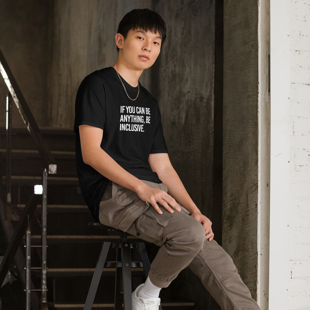 This is the photo of a black Be Inclusive Basic Tee. On the front of the t-shirt, the phrase, 'If you can be anything, be inclusive,' is printed in white capital letters. A man is sitting on a stool wearing the tee.