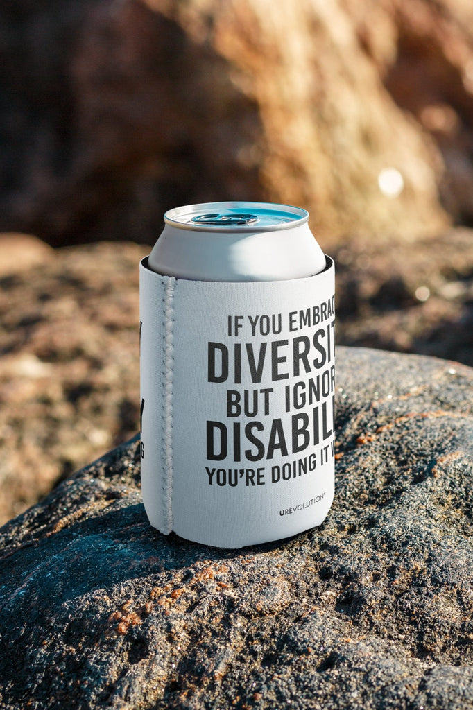 A white 12 oz Embrace Diversity Koozie with the phrase, 'If you embrace diversity but ignore disability, you're doing it wrong.' printed in black on the front and back of the koozie. The koozi is placed on a rock on the beach.
