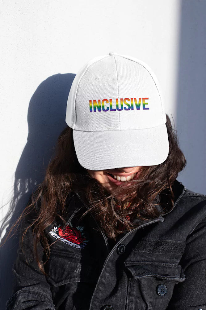 Woman wearing a white gay pride hat with rainbow-colored "INCLUSIVE" text, smiling in the sunlight and dressed in a black denim jacket—celebrating LGBTQ+ pride and inclusion.
