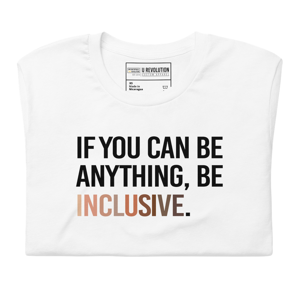 A photo of a white Be Inclusive t-shirt promoting racial diversity. On the front of the inclusive t-shirt is the phrase: "If you can be anything, be inclusive," in black upper case letters. The text is all black, except the word "Inclusive," which is in different skin colors, with the lightest color on the left and the darkest on the right. The shirt is folded.