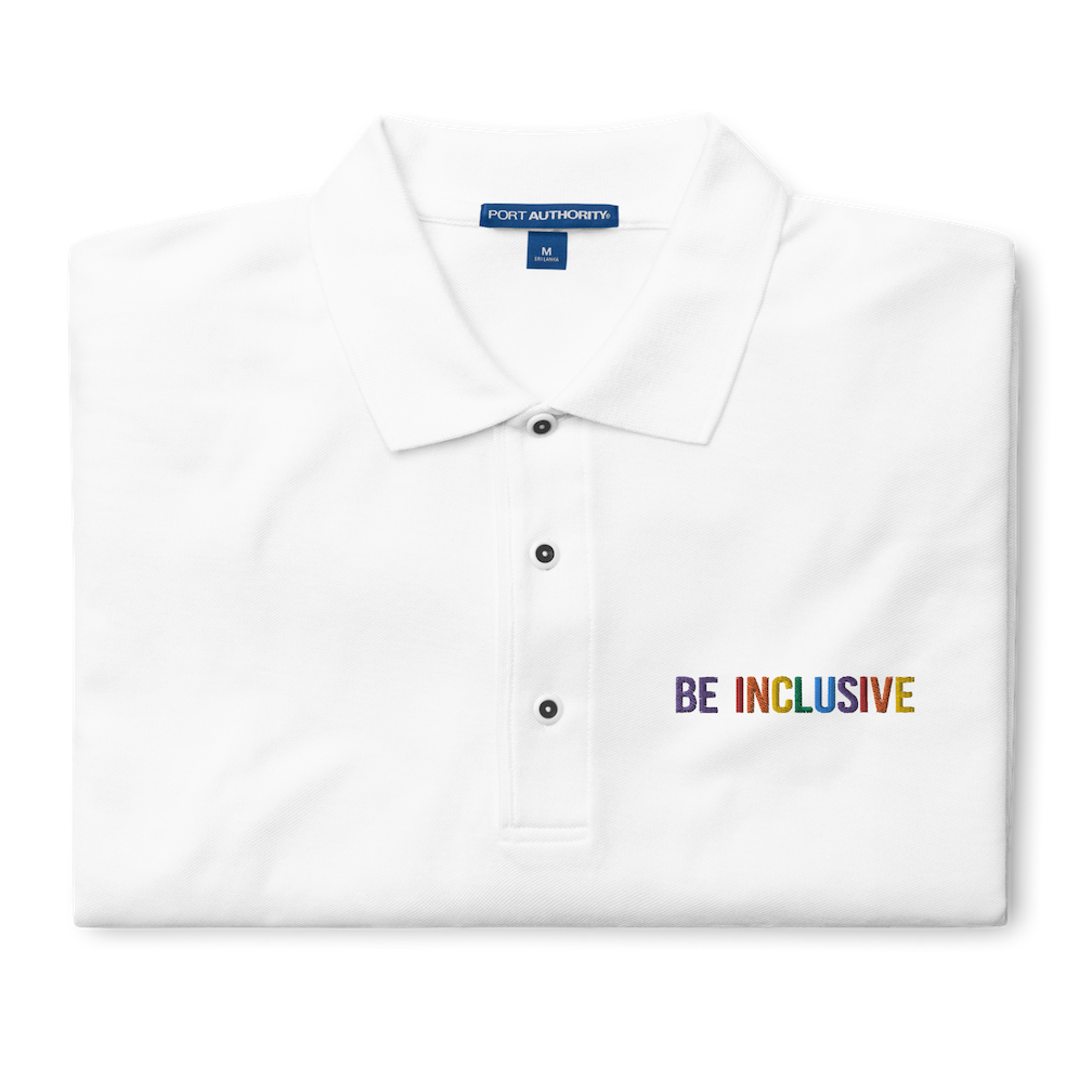 This is a photo of a premium white Rainbow Polo Shirt. On the polo's top right panel is the phrase, Be Inclusive, embroidered in red, orange, yellow, green, indigo, and violet. The word 'be' is only in violet. The polo is folded neatly.