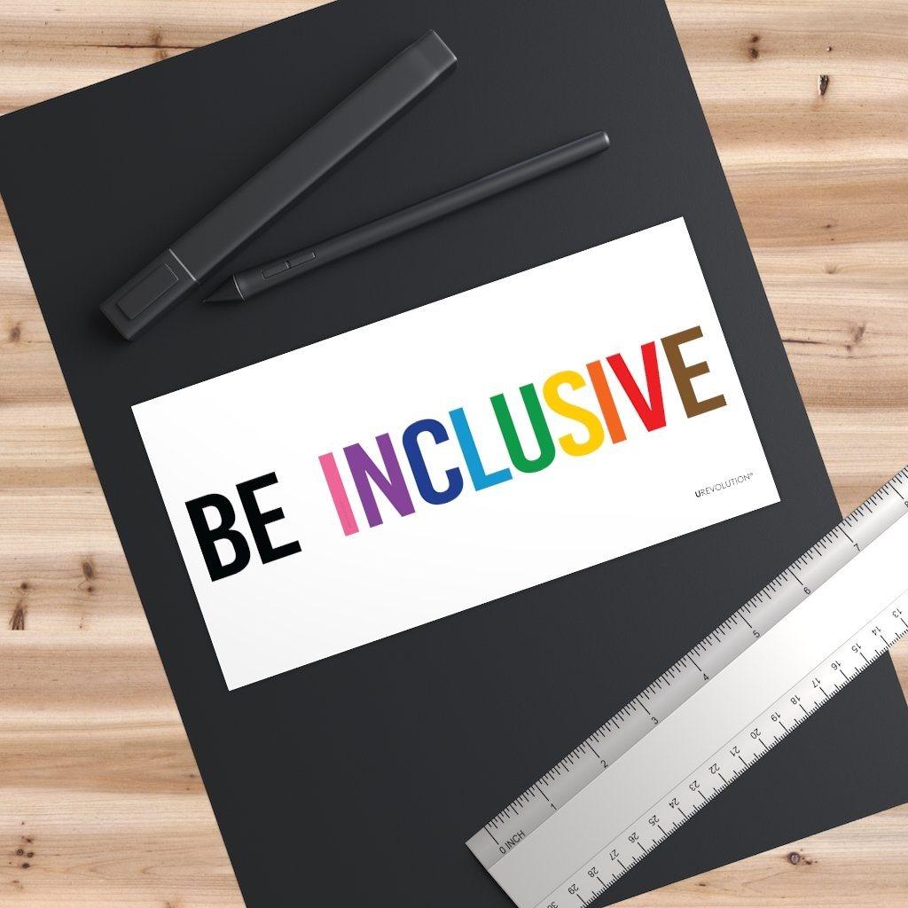 A photo of an waterproof inclusive bumper sticker on a black desk pad. The words "Be Inclusive" are printed on a white background in upper case letters. The word "be" is in black, and the word "inclusive" is in the rainbow colors. In small upper case letters, in the bottom corner of the sticker, is the word URevolution