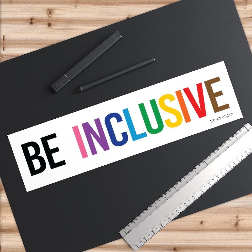 A photo of an inclusive bumper sticker on a black desk pad. The words "Be Inclusive" are printed on a white background in upper case letters. The word "be" is in black, with word "inclusive" is in the colors of the rainbow. In small upper case letters, in the bottom corner of the sticker, is the word URevolution.