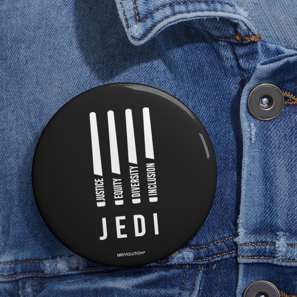 A photo of a JEDI pin by URevolution. In the middle of the black JEDI pin are four white lightsabers. At the bottom of each saber representing the saber handle is one word: Justice Equity Diversity Inclusion. Beneath the sabers is the acronym JEDI. The 3" JEDI pin is displayed on a blue denim jacket.