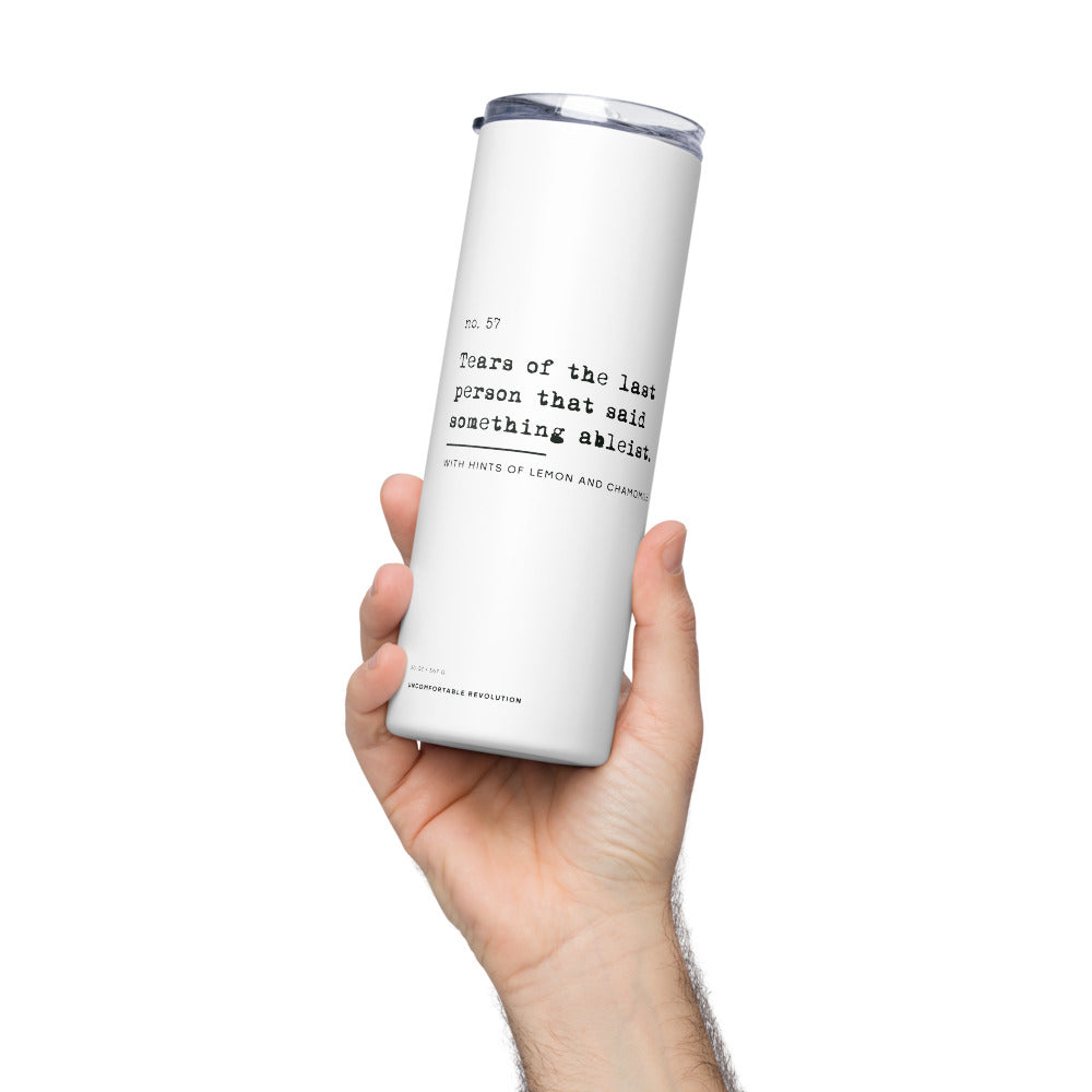 The Ableist Tears Stainless Steel Travel Mug is white, with black text that reads: "no. 57. Tears of the last person that said something ableist. With hints of lemon and chamomile." At the base of the mug is the text: 20oz / 567 gr Uncomfortable Revolution. The mug has a plastic lid and metal straw. The mug is held in a person's hand.