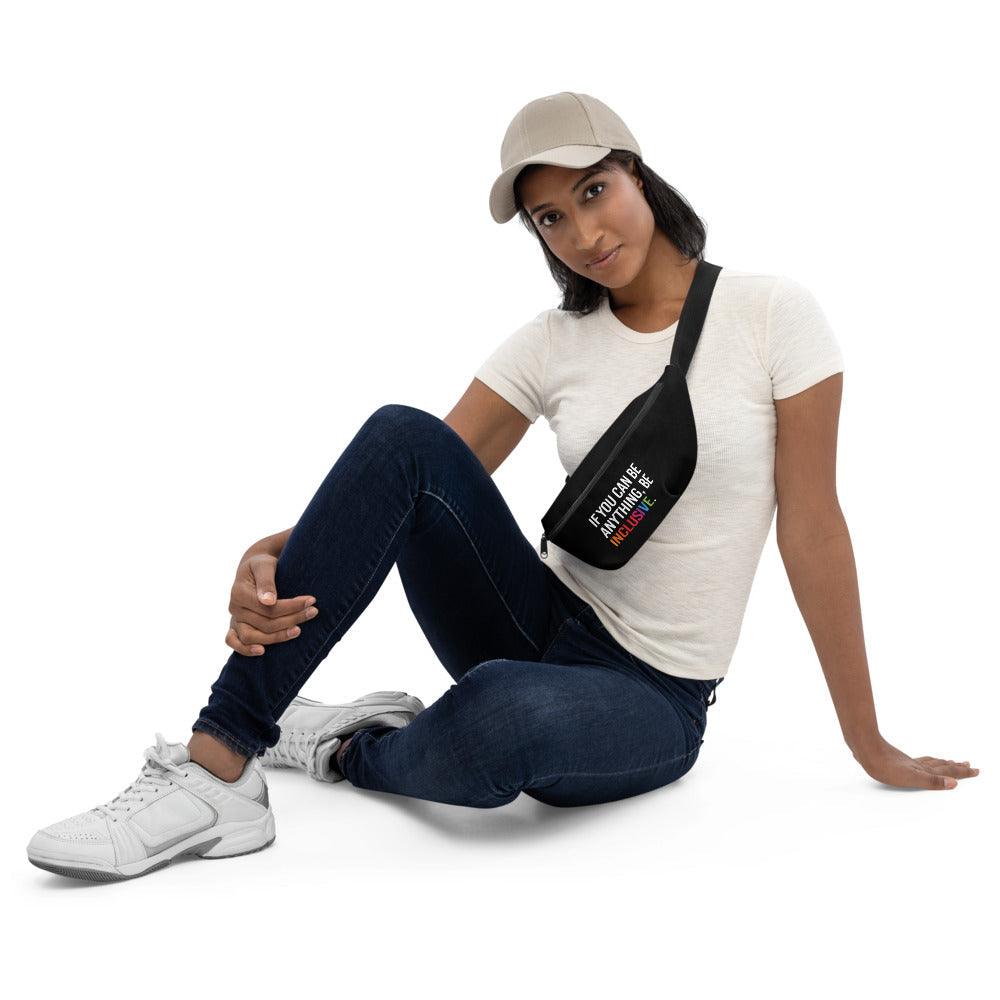 This is a photo of a black Be Inclusive waist bag. On the front of the waist bag is the phrase, 'If you can be anything, be inclusive,' in white upper case letters. The word inclusive is written in the colors of the rainbow. The bag is worn by a model down with the waist bag across their chest.