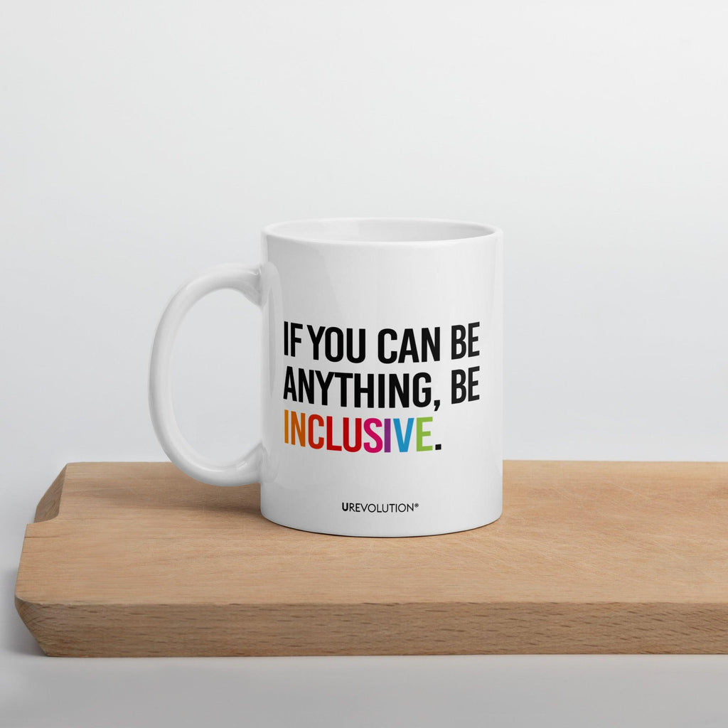 11oz Be Inclusive mug. On the right-hand side of the mug, facing the screen, is the phrase, 'If you can be anything, be inclusive,' printed in large black upper case letters. The word 'inclusive' is in the color of the rainbow. At the bottom edge of the inclusive mug is the word, URevolution, printed in small black upper case letters. The inclusive mug is sitting on a wooded cutting board.