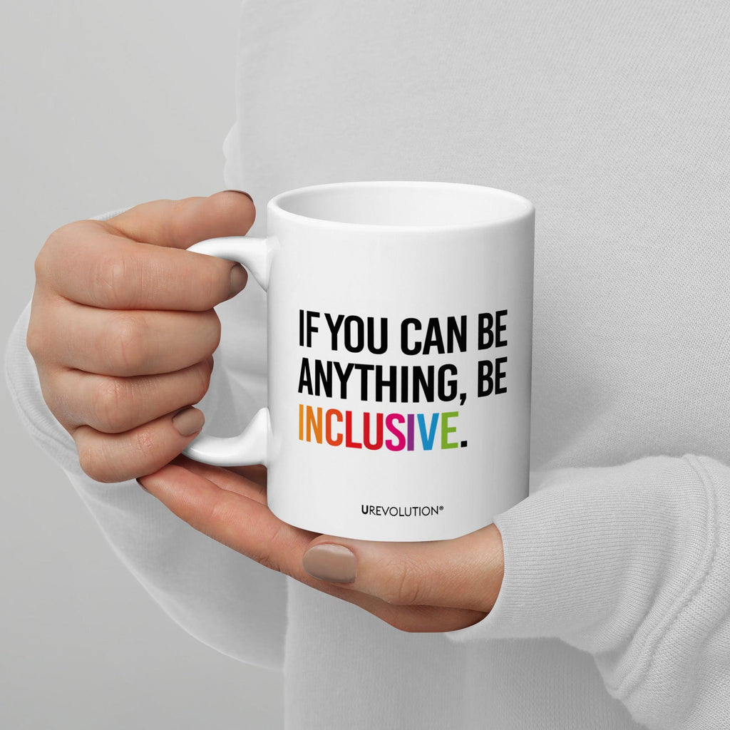 11oz Be Inclusive mug. On the right-hand side of the mug, facing the screen, is the phrase, 'If you can be anything, be inclusive,' printed in large black upper case letters. The word 'inclusive' is in the color of the rainbow. At the bottom edge of the inclusive mug is the word, URevolution, printed in small black upper case letters. A person is holding the mug in their right hand.