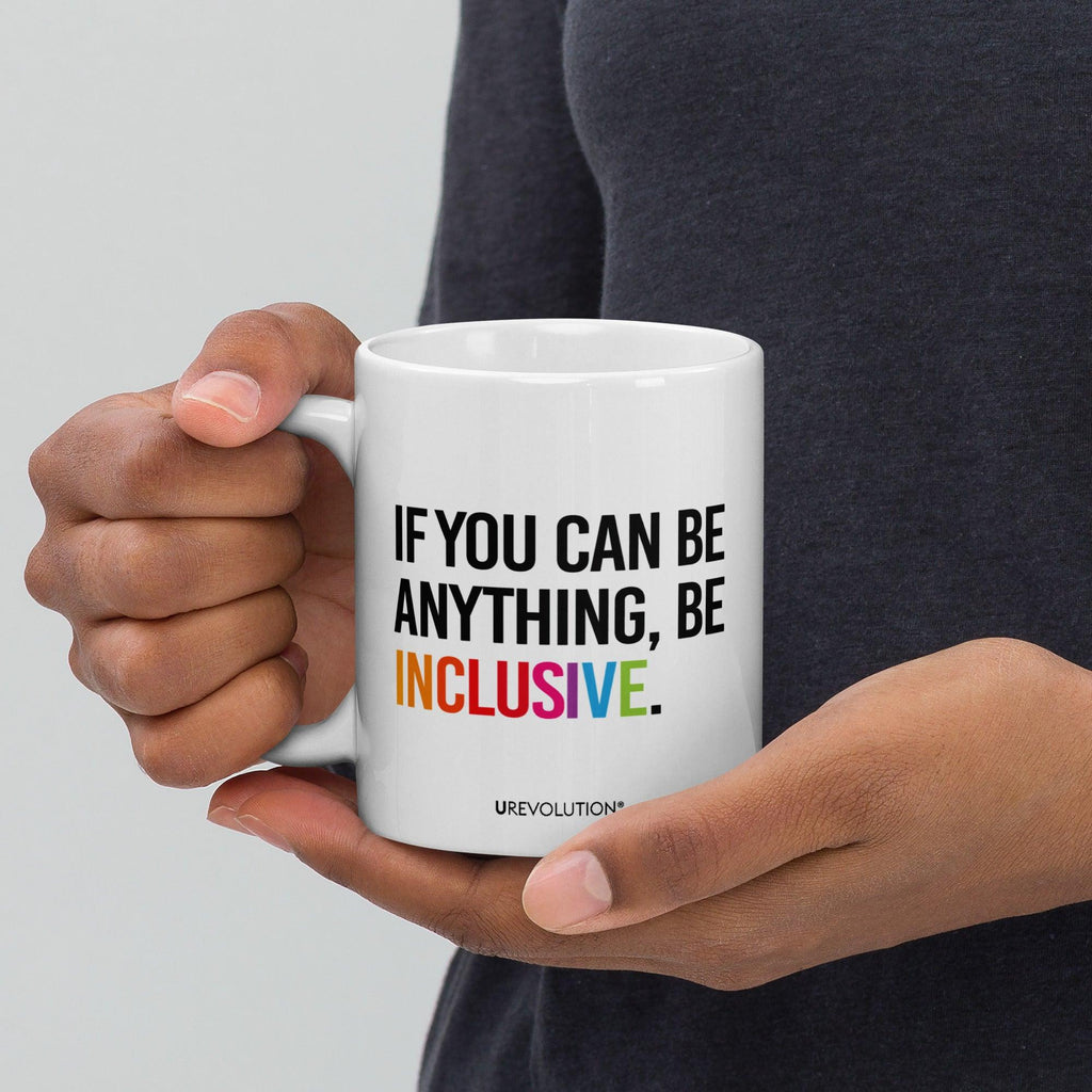 11 oz Be Inclusive mug. On the right-hand side of the mug, facing the screen, is the phrase, 'If you can be anything, be inclusive,' printed in large black upper case letters. The word 'inclusive' is in the color of the rainbow. At the bottom edge of the inclusive mug is the word, URevolution, printed in small black upper case letters. A person is holding the mug in their right hand.
