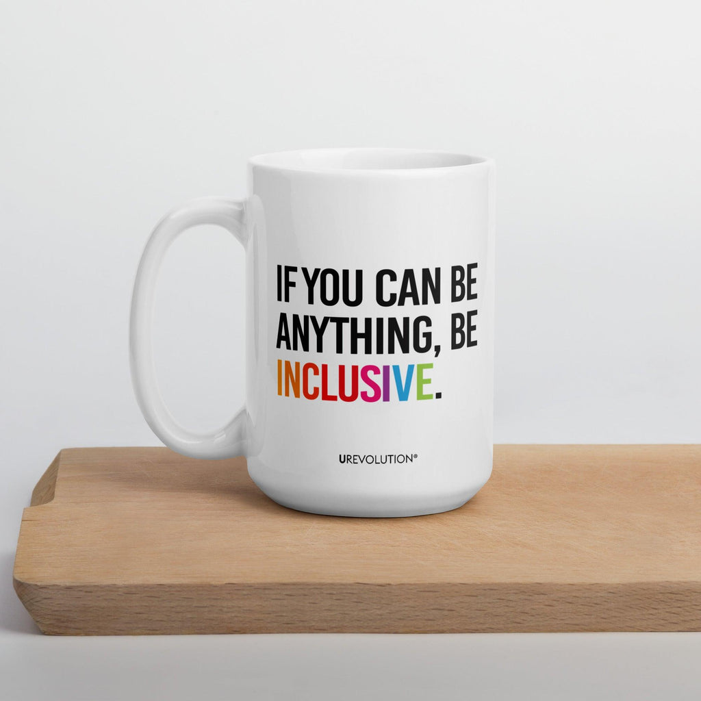 15oz Be Inclusive mug. On the right-hand side of the mug, facing the screen, is the phrase, 'If you can be anything, be inclusive,' printed in large black upper case letters. The word 'inclusive' is in the color of the rainbow. At the bottom edge of the inclusive mug is the word, URevolution, printed in small black upper case letters. The mug is sitting on a wooden cutting board.
