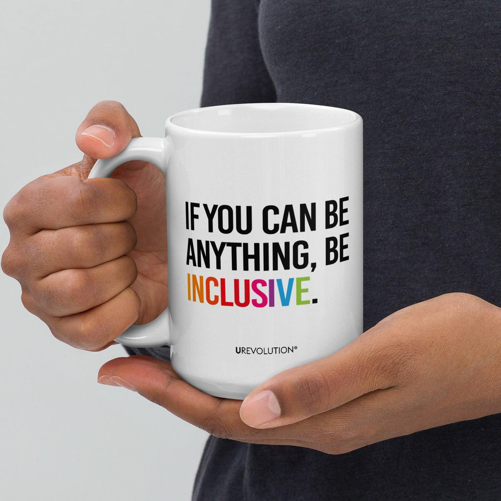 15oz Be Inclusive mug. On the right-hand side of the mug, facing the screen, is the phrase, 'If you can be anything, be inclusive,' printed in large black upper case letters. The word 'inclusive' is in the color of the rainbow. At the bottom edge of the inclusive mug is the word, URevolution, printed in small black upper case letters. A person is holding the mug in their right hand.