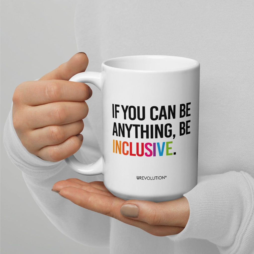 15oz Be Inclusive mug. On the right-hand side of the mug, facing the screen, is the phrase, 'If you can be anything, be inclusive,' printed in large black upper case letters. The word 'inclusive' is in the color of the rainbow. At the bottom edge of the inclusive mug is the word, URevolution, printed in small black upper case letters. A person is holding the mug in their right hand.