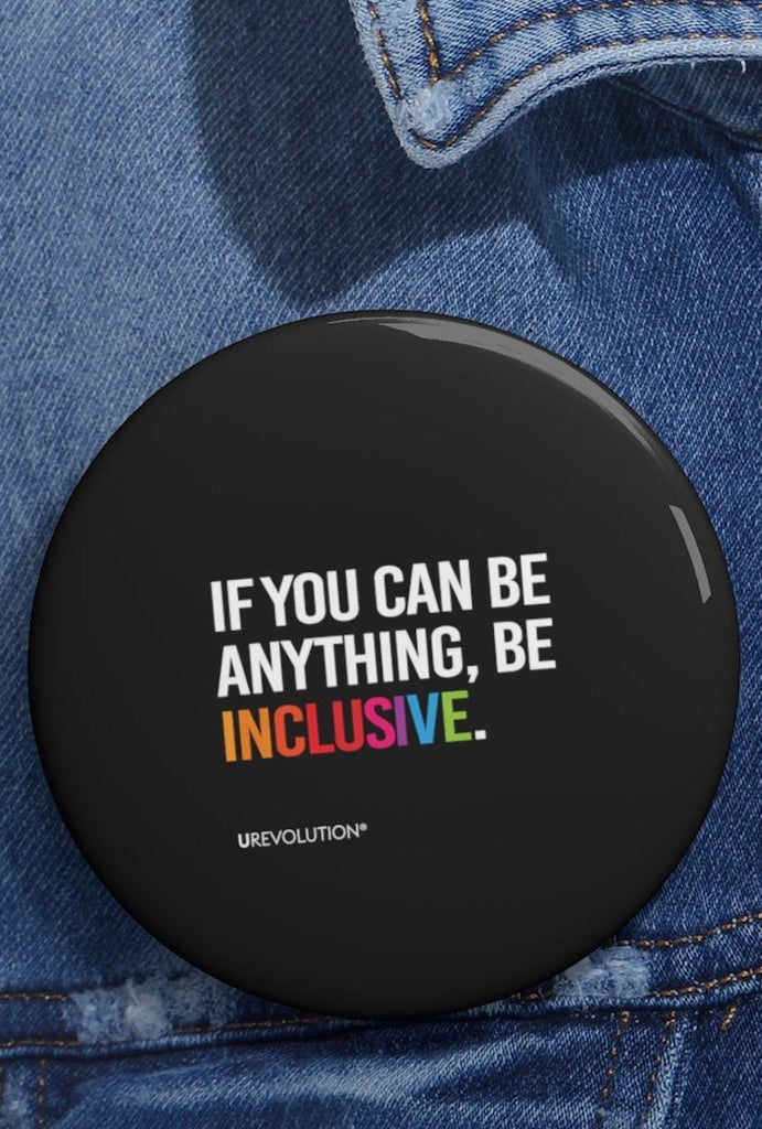 A Be Inclusive pin button. The phrase, "if you can be anything, be inclusive," is printed in upper white case letters against a black background. The word inclusive is in the colors of the rainbow.