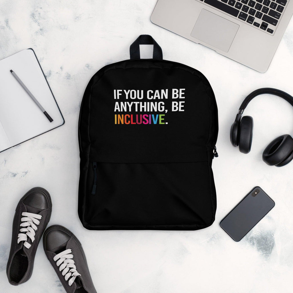 This is a photo of URevolution's black Be Inclusive backpack. In the top of half of the backpack, the phrase, 'If you can be anything, be inclusive,' is printed in white upper case letters. The rest of the backpack is black. The word 'inclusive' is in the color of the rainbow.  The backpack is in the middle of table. Around the backup is a laptop, headphones, iPhone, and a notepad.