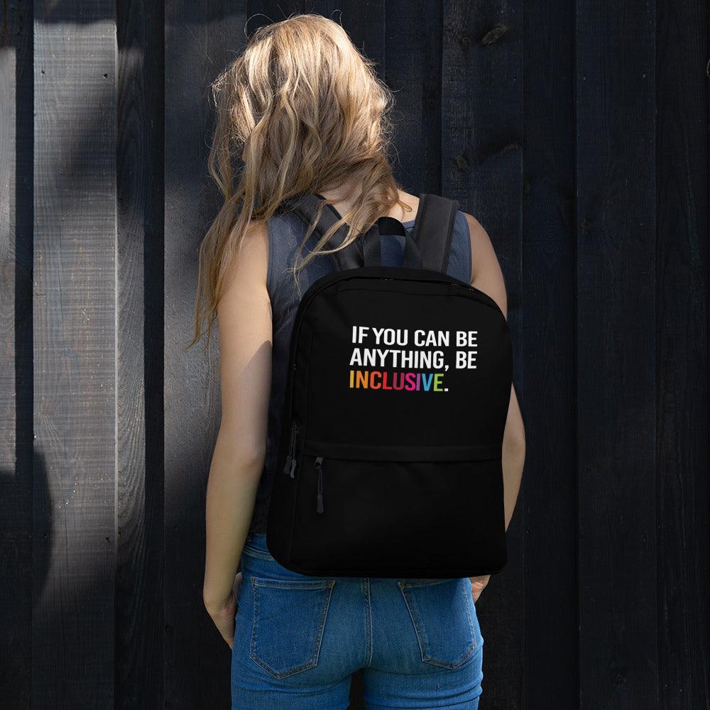 This is a photo of URevolution's black Be Inclusive backpack. In the top of half of the backpack, the phrase, 'If you can be anything, be inclusive,' is printed in white upper case letters. The rest of the backpack is black. The word 'inclusive' is in the color of the rainbow. The backpack is being worn on the back of model with long fawn hair, wearing a navy tank top and blue jeans. The model is standing outside in front of black wooden fence in the sunlight.
