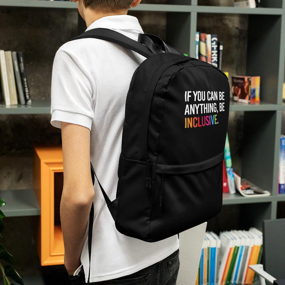 This is a photo of URevolution's black Be Inclusive backpack. In the top of half of the backpack, the phrase, 'If you can be anything, be inclusive,' is printed in white upper case letters. The rest of the backpack is black. The word 'inclusive' is in the color of the rainbow. The backpack is work by a teenager wearing a white polo shirt and black jeans standing in a school library.