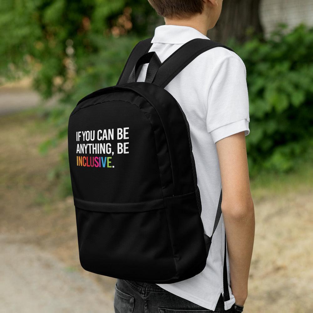 This is a photo of URevolution's black Be Inclusive backpack. In the top of half of the backpack, the phrase, 'If you can be anything, be inclusive,' is printed in white upper case letters. The rest of the backpack is black. The word 'inclusive' is in the color of the rainbow. The backpack is work by a teenager wearing a white polo shirt and black jeans standing outside in a park.