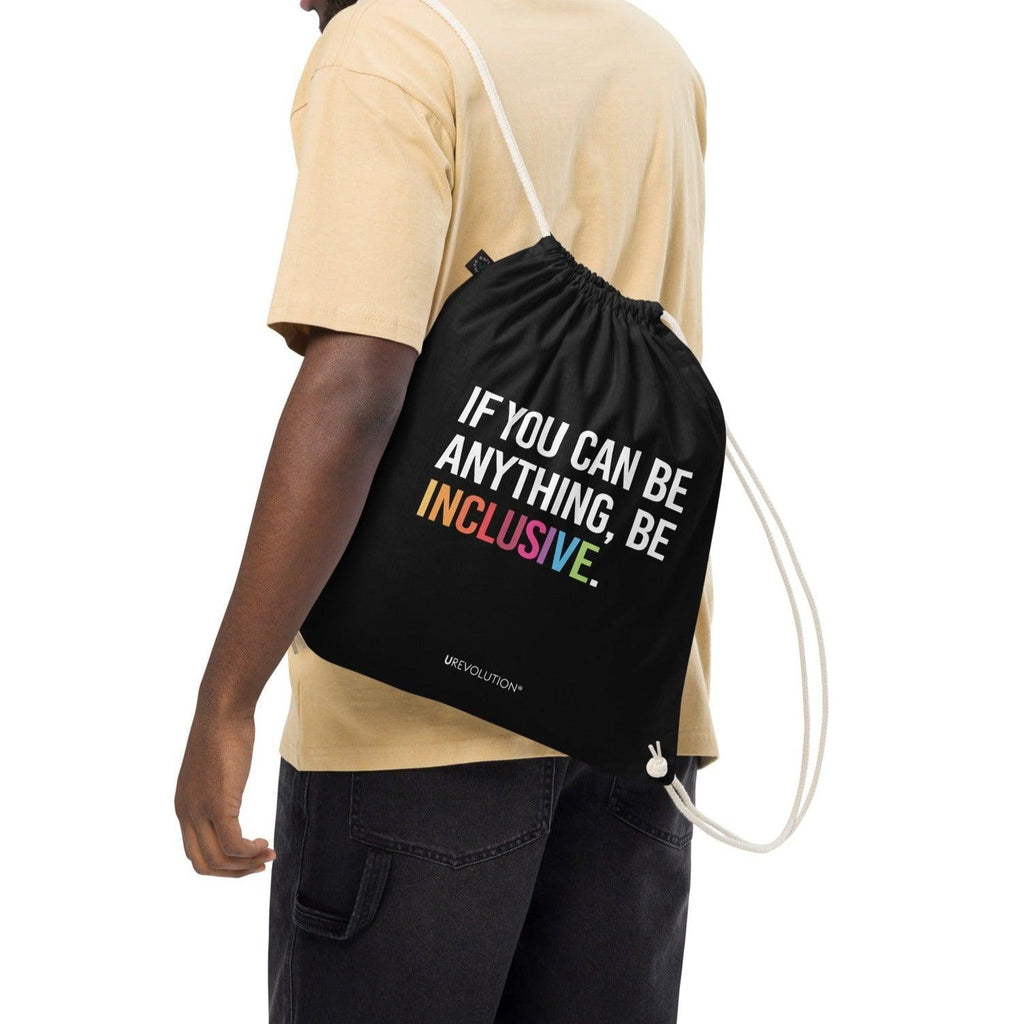 This is a photo of URevolution's black Be Inclusive organic cotton drawstring bag. In the middle of the drawstring bag, printed in large white upper case letters, is the phrase, 'If you can be anything, be inclusive.' The word 'inclusive' is in the color of the rainbow. At the bottom edge of the bag is the word, URevolution, printed in small white upper case letters. The bag's cotton drawstrings are pulling the top of the bag closed. The bag is slung over the shoulders of a model.