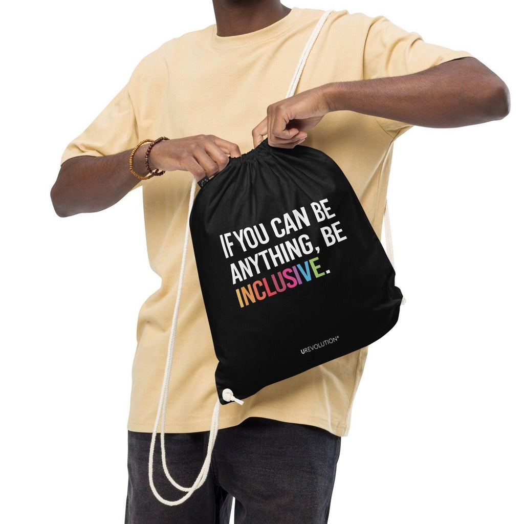 This is a photo of URevolution's black Be Inclusive organic cotton drawstring bag. In the middle of the drawstring bag, printed in large white upper case letters, is the phrase, 'If you can be anything, be inclusive.' The word 'inclusive' is in the color of the rainbow. At the bottom edge of the bag is the word, URevolution, printed in small white upper case letters. The bag's cotton drawstrings are pulling the top of the bag closed. The bag is slung over the shoulders of a model.