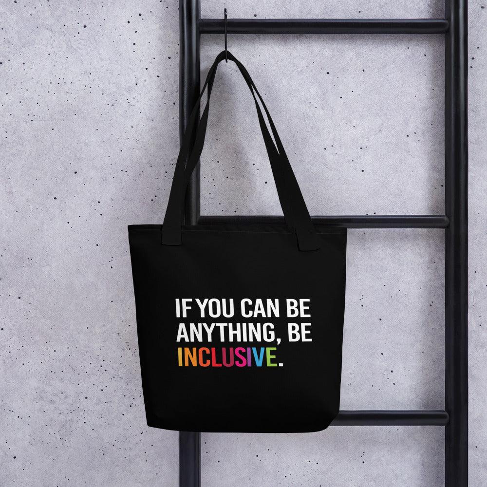 Inclusive Tote Bag. The black tote has the phrase, 'If you can be anything, Be Inclusive,' printed in white. The word inclusive is in the colors of the rainbow. The inclusive tote is hung on a ladder.