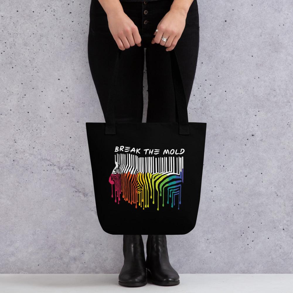 An image of a black tote bag against a plain background. In the middle of the bag is a graphic of zebra in the silhouette of a barcode. The top half of the zebra are vertical white stripes, which then change just below the back into the colours of the rainbow. The bottom of each stripe has a paint drip mark. Just above the zebra's back is the phrase, in upper case, 'break the mold.'