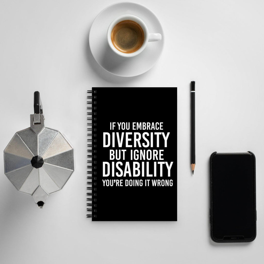 Front cover of URevolution's spiral-bound black Embrace Diversity notebook. The cover features URevolution's original Embrace Diversity phrase printed in white on a black background: "If you embrace diversity but ignore disability, you're doing it wrong." The embrace diversity notebook is placed in the middle of the table.