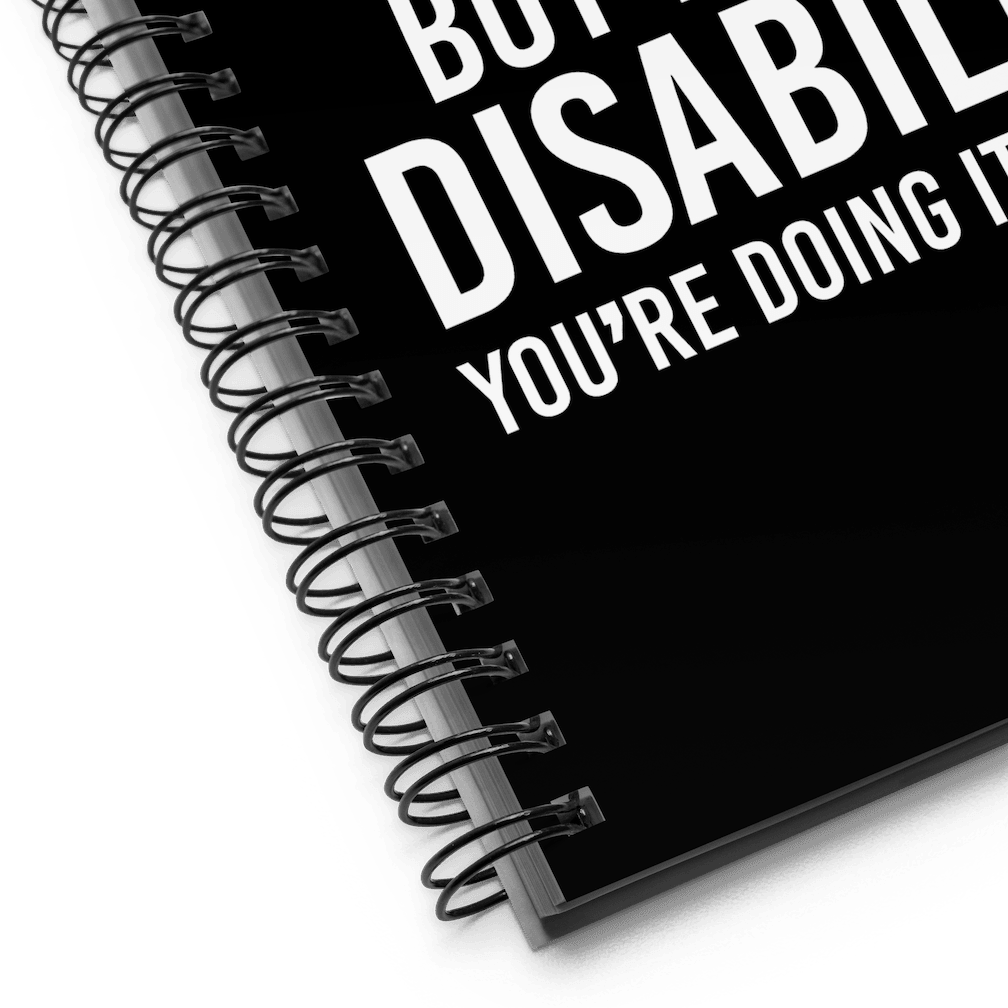Front cover of URevolution's spiral-bound black Embrace Diversity notebook. The cover features URevolution's original Embrace Diversity phrase printed in white on a black background: "If you embrace diversity but ignore disability, you're doing it wrong." The photo zooms into the bottom left hand corner of the notebook.
