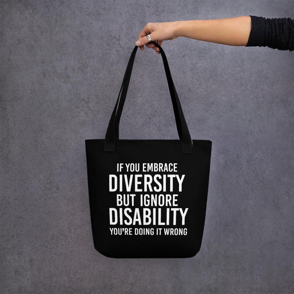 Photo of a black Embrace Diversity Tote with the phrase, "If you embrace diversity, but ignore disability, you're doing it wrong," printed on it in white upper case letters. The disability inclusion tote is held in the air by a person's arm.