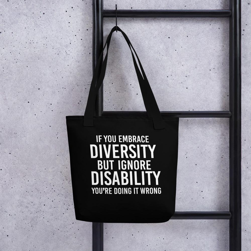 Photo of a black Embrace Diversity Tote with the phrase, "If you embrace diversity, but ignore disability, you're doing it wrong," printed on it in white upper case letters. The disability inclusion tote is hing on a black ladder.