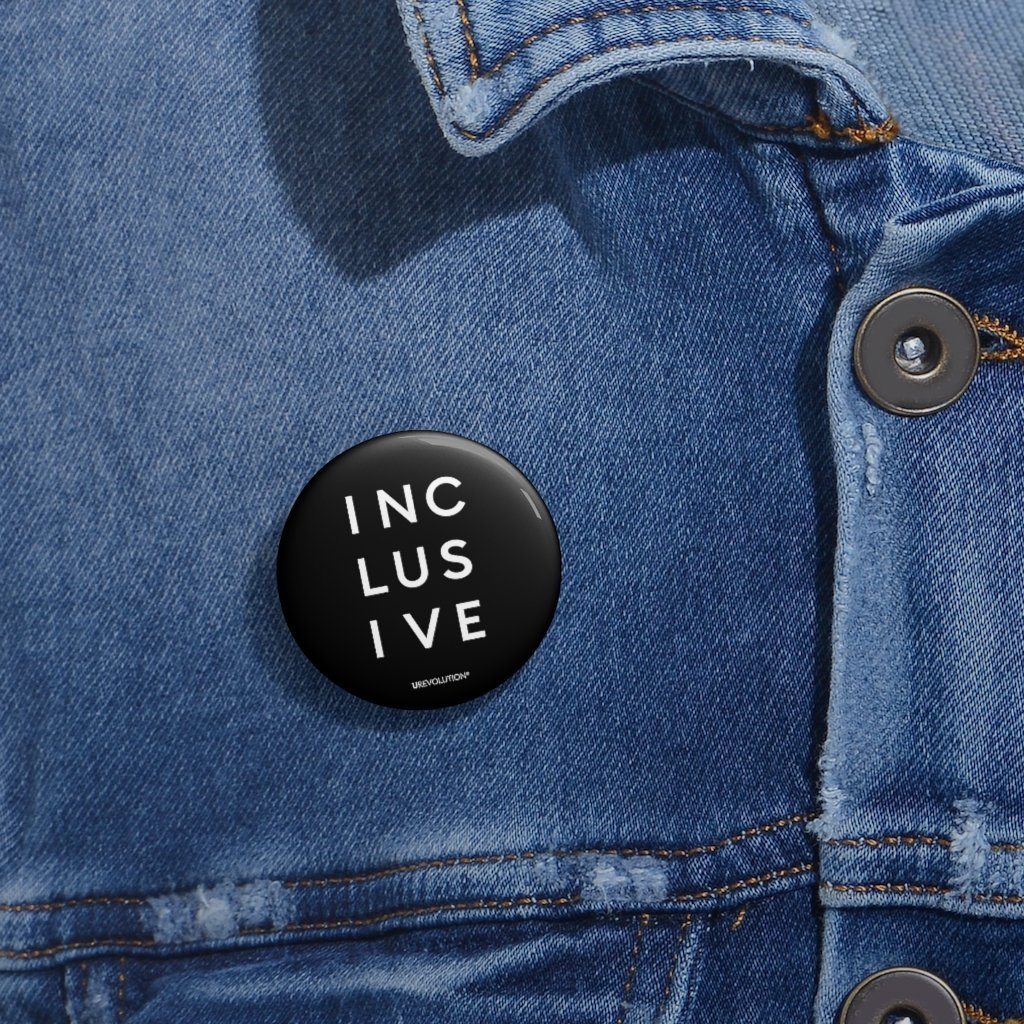 1.25" black inclusive pin button. On the front of the inclusive pin button is the word inclusive. The word inclusive is printed in black over three lines, with three letters on each line: INC LUS IVE. On the bottom edge of the pin, beneath the word inclusive, is the word URevolution in a small discrete font. horizontal black line. The pin is positioned on the left-hand side of a blue denim jacket beneath the lapel.
