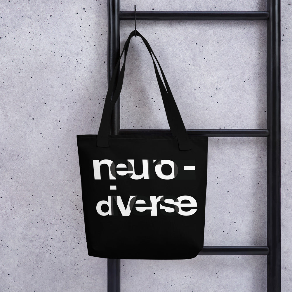 In the middle of the neurodiverse black tote bag, the word NEURODIVERSE is printed in large alternating black and white letters, taking up about two-thirds of the tote bag surface. The tote bag is hanging on a black ladder leaning against a grey concrete wall.