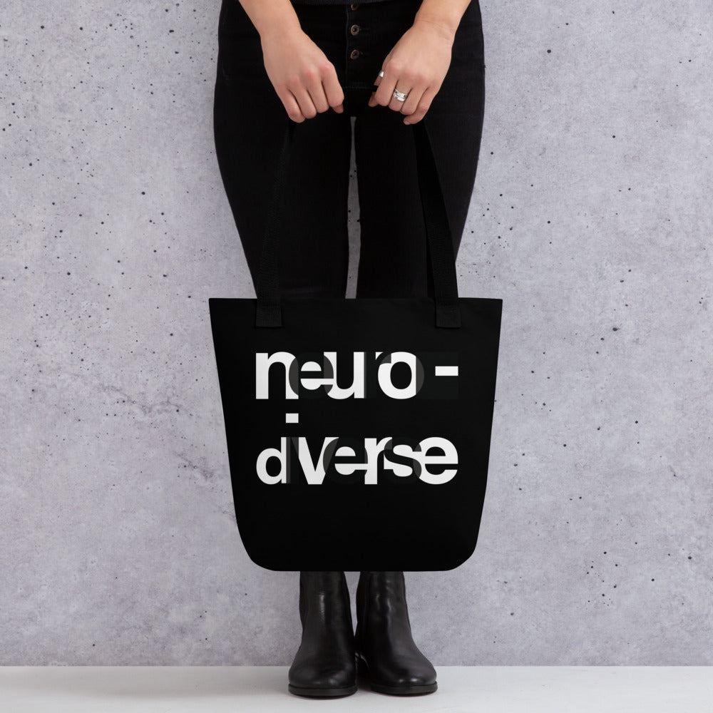 In the middle of the neurodiverse black tote bag, the word NEURODIVERSE is printed in large alternating black and white letters, taking up about two-thirds of the tote bag surface. The tote bag is being held in front of a model only their torso is visible. 