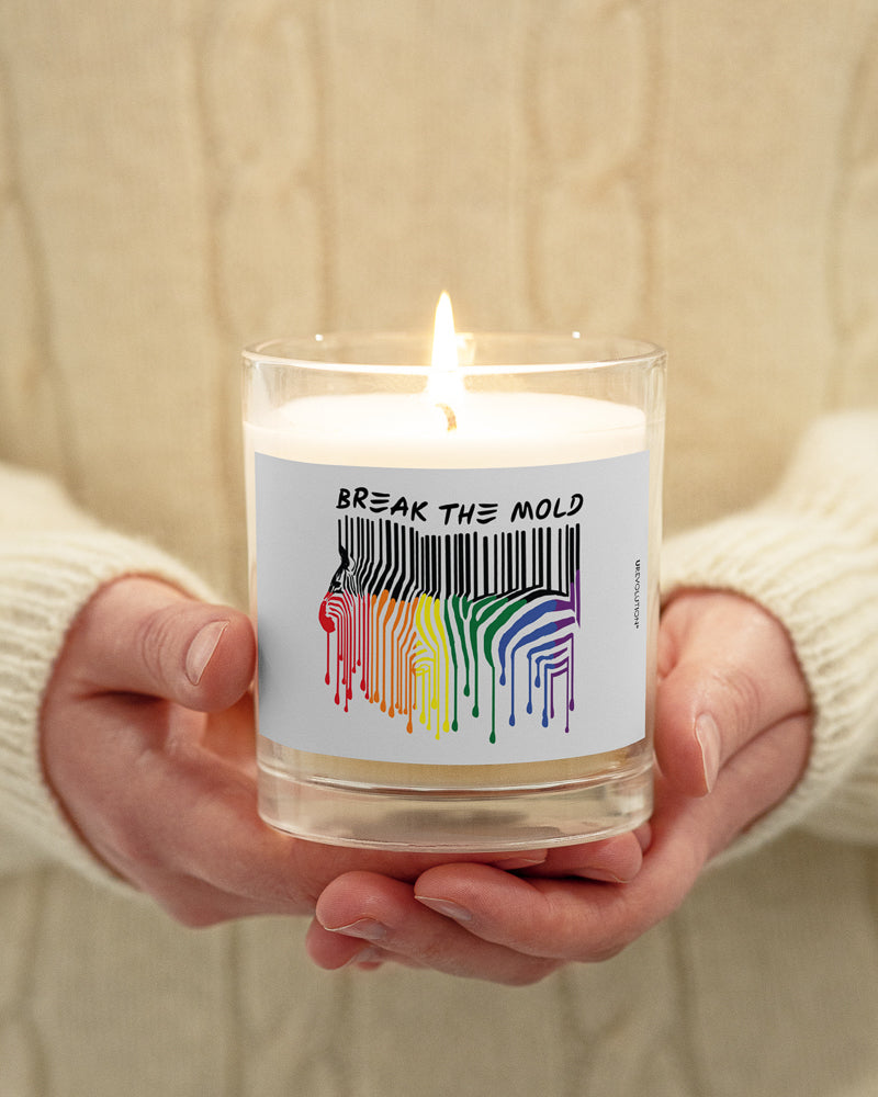 Break The Mold soy wax candle. In the middle of the candle is a zebra graphic in the silhouette of a barcode. The top half of the zebra are vertical black stripes, which change just below the back into the colors of the rainbow. The bottom of each strip has a paint drip mark. Above the zebra's back is the phrase, in upper case, 'break the mold.'  A light Break The Mold candle is held in a person's hands.