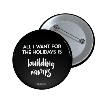 A photo of a round black Building Ramps pin. In the middle of the equal pay pin phrase, 'All I want for the holidays is,' printed in large black upper case letters, with the words, 'building ramps,' printed directly beneath that phrase in black elegant cursive font. 