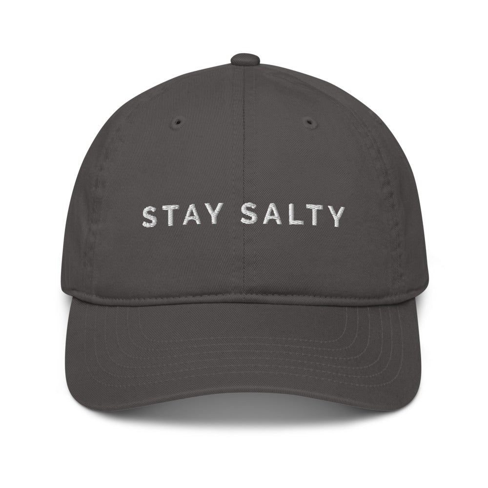 A photo of a charcoal-colored organic Stay Salty cap lying flat against a plain background. On the front panel of the cap the phrase, 'Stay Salty,' is embroidered on it in white upper case letters. 