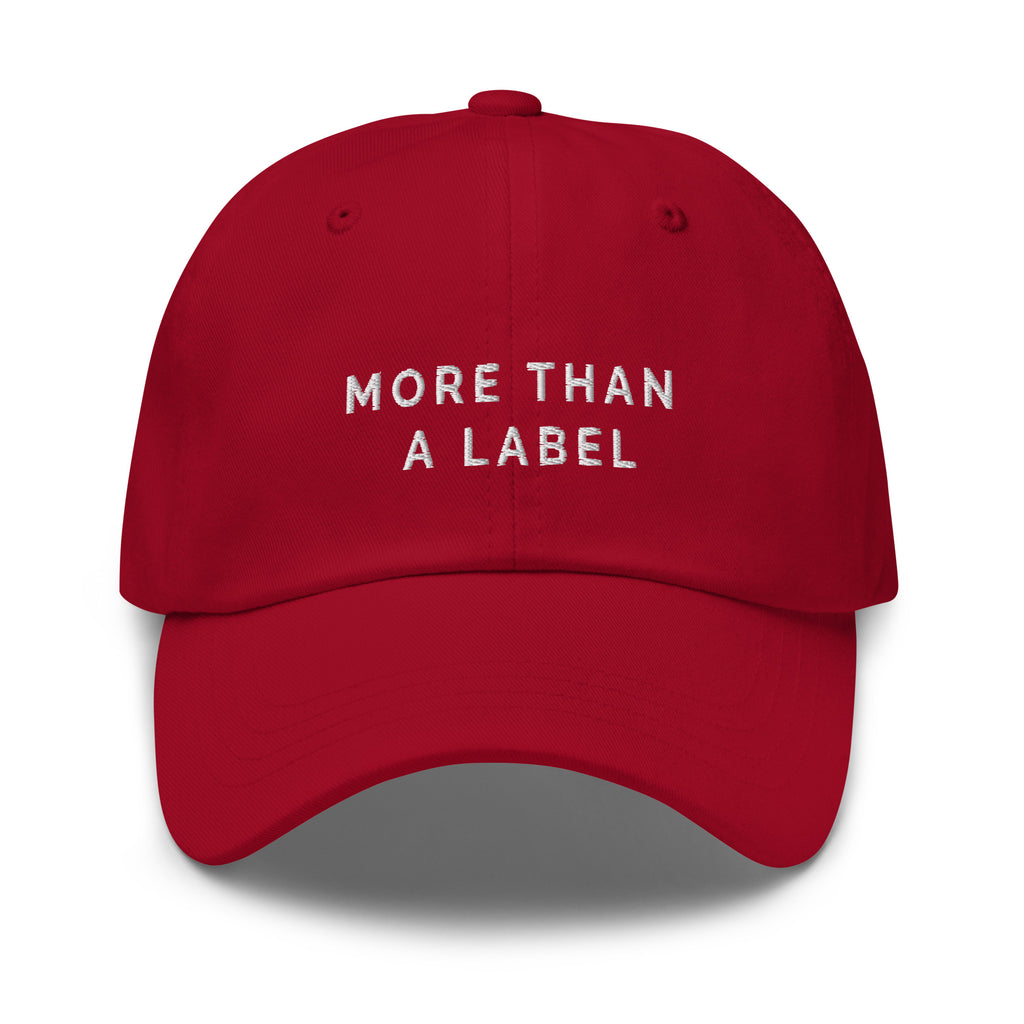 A cranberry More Than A Label cap. In the middle of the cap is the phrase, 'More Than A Label,' embroidered in white upper case letters.