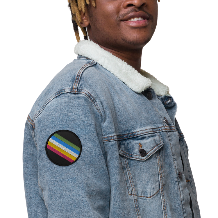 This is an image of an embroidered Disability Pride Flag Patch. In the middle of the black patch, there are five diagonal stripes in this order: red, yellow, white, blue, and green (when looking from the bottom up).