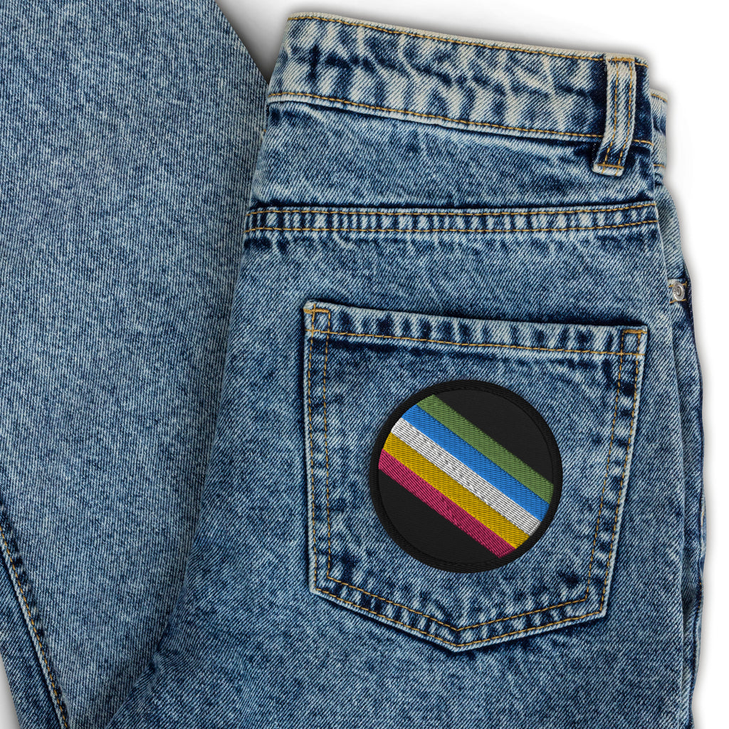 This is an image of an embroidered Disability Pride Flag Patch. In the middle of the black patch, there are five diagonal stripes in this order: red, yellow, white, blue, and green (when looking from the bottom up).
