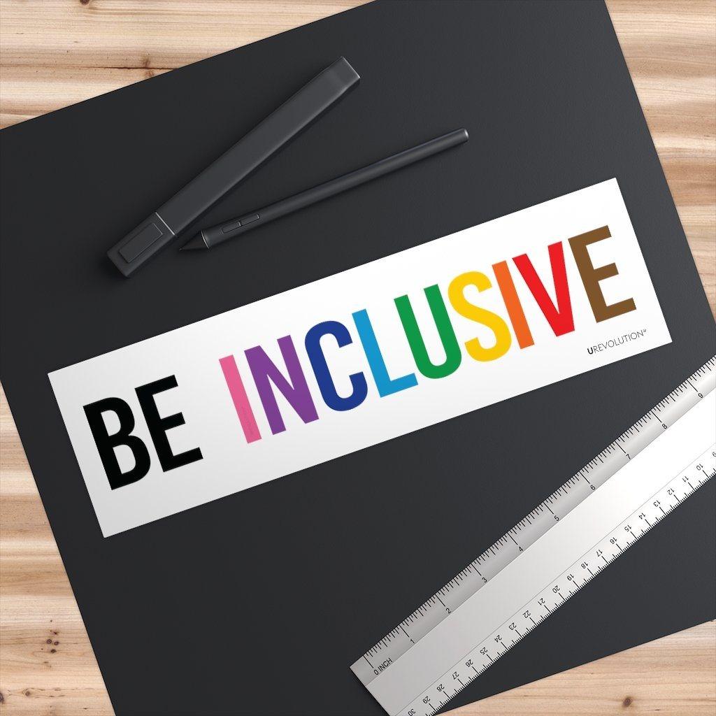A photo of an waterproof inclusive bumper sticker on a black desk pad. The words "Be Inclusive" are printed on a white background in upper case letters. The word "be" is in black, and the word "inclusive" is in the rainbow colors. In small upper case letters, in the bottom corner of the sticker, is the word URevolution