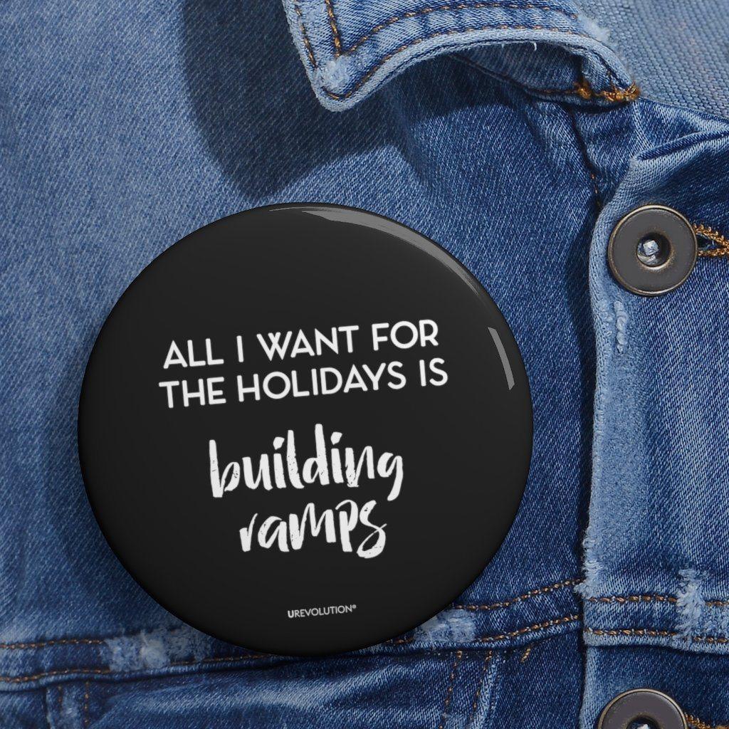 A close-up photo of a round black Building Ramps pin on a blue denim jacket. In the middle of the equal pay pin phrase, 'All I want for the holidays is,' printed in large black upper case letters, with the words, 'building ramps,' printed directly beneath that phrase in black elegant cursive font.