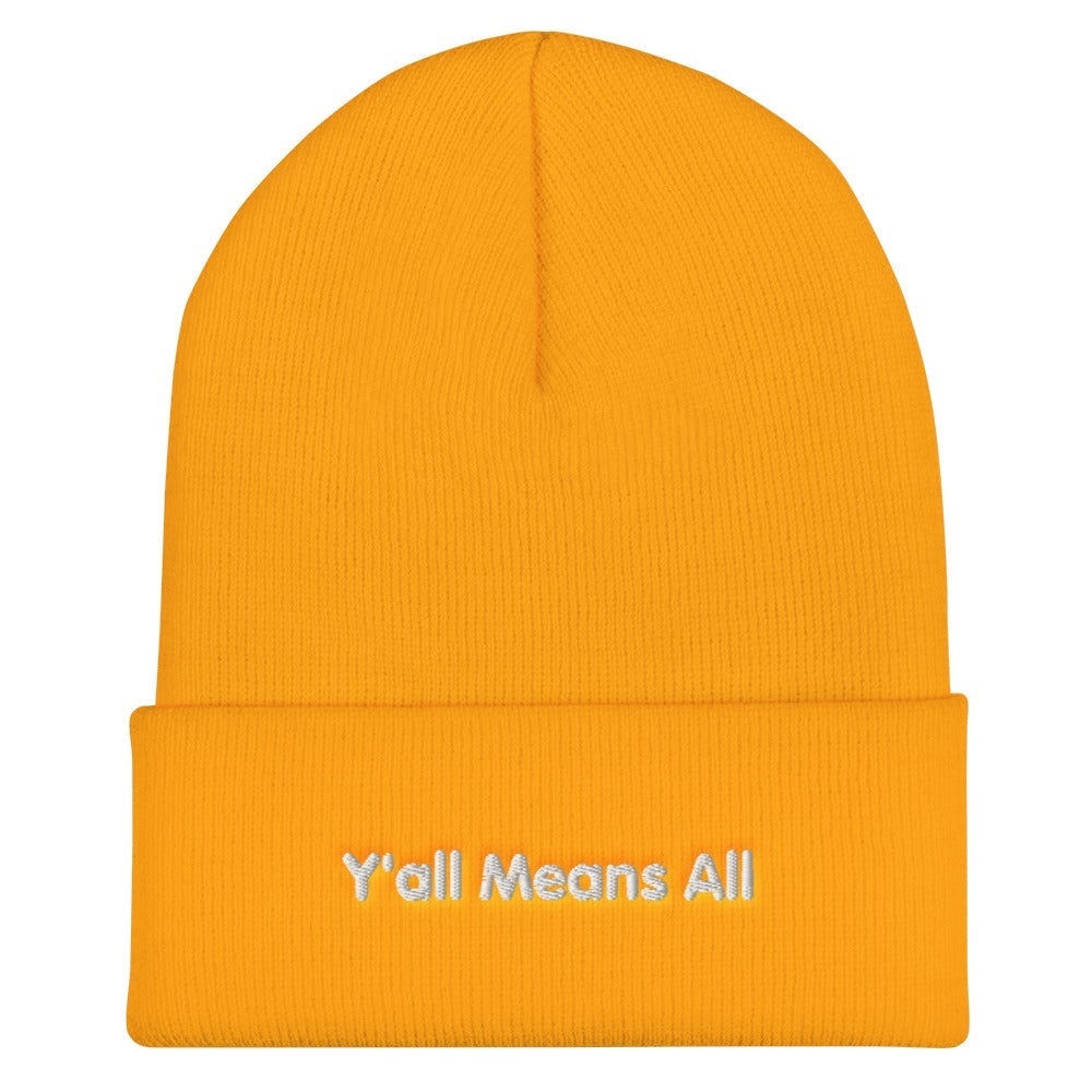 This is a photo of a gold-colored Y'all Means All beanie. On the front of the beanie the phrase, 'Y'all Means All,' is embroidered on it in white title case letters. 