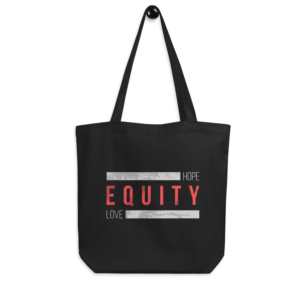 A black HELO Equity Organic Tote Bag. The word 'Equity' in upper case red letters is in the middle of the tote bag. Above and below the word are two thick distressed grey rectangle blocks with the word HOPE printed on the top right-hand side, and the word LOVE on the bottom right side of the block. The equity tote is hanging on a wall hook.