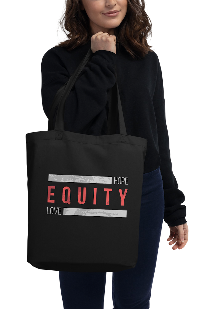 A black HELO Equity Organic Tote Bag. The word 'Equity' in upper case red letters is in the middle of the tote bag. Above and below the word are two thick distressed grey rectangle blocks with the word HOPE printed on the top right-hand side, and the word LOVE on the bottom right side of the block. The bag is being held by a model in front of their body.