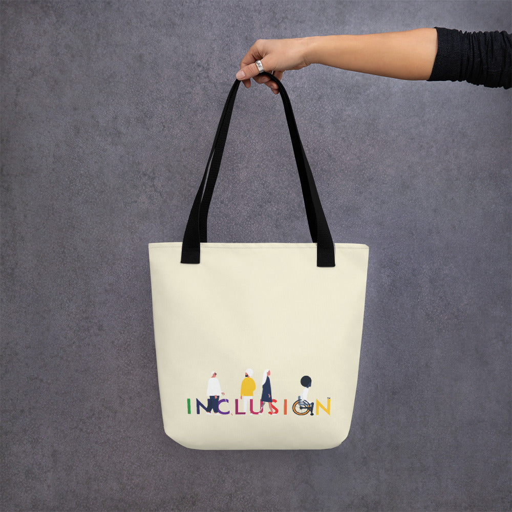 A disability inclusion tote bag has the word INCLUSION printed in rainbow-like colors on one side. The inclusion tote is canvas color with black straps. Four diverse disabled people have incorporated into the phrase INCLUSION: a blind person, a person with an invisible illness, a disabled person with a prosthetic limb, and a disabled person in a wheelchair. The bag is being held in a persons hand.