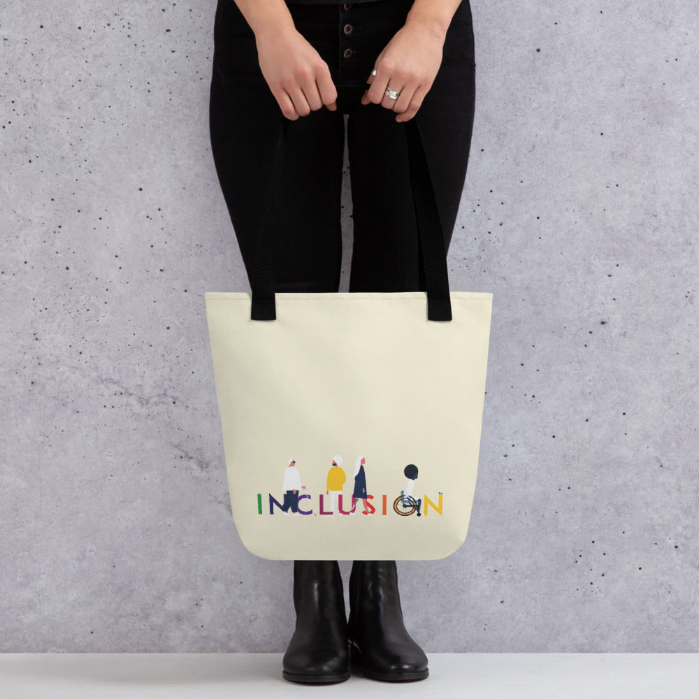 A disability inclusion tote bag has the word INCLUSION printed in rainbow-like colors on one side. The inclusion tote is canvas color with black straps. Four diverse disabled people have incorporated into the phrase INCLUSION: a blind person, a person with an invisible illness, a disabled person with a prosthetic limb, and a disabled person in a wheelchair.