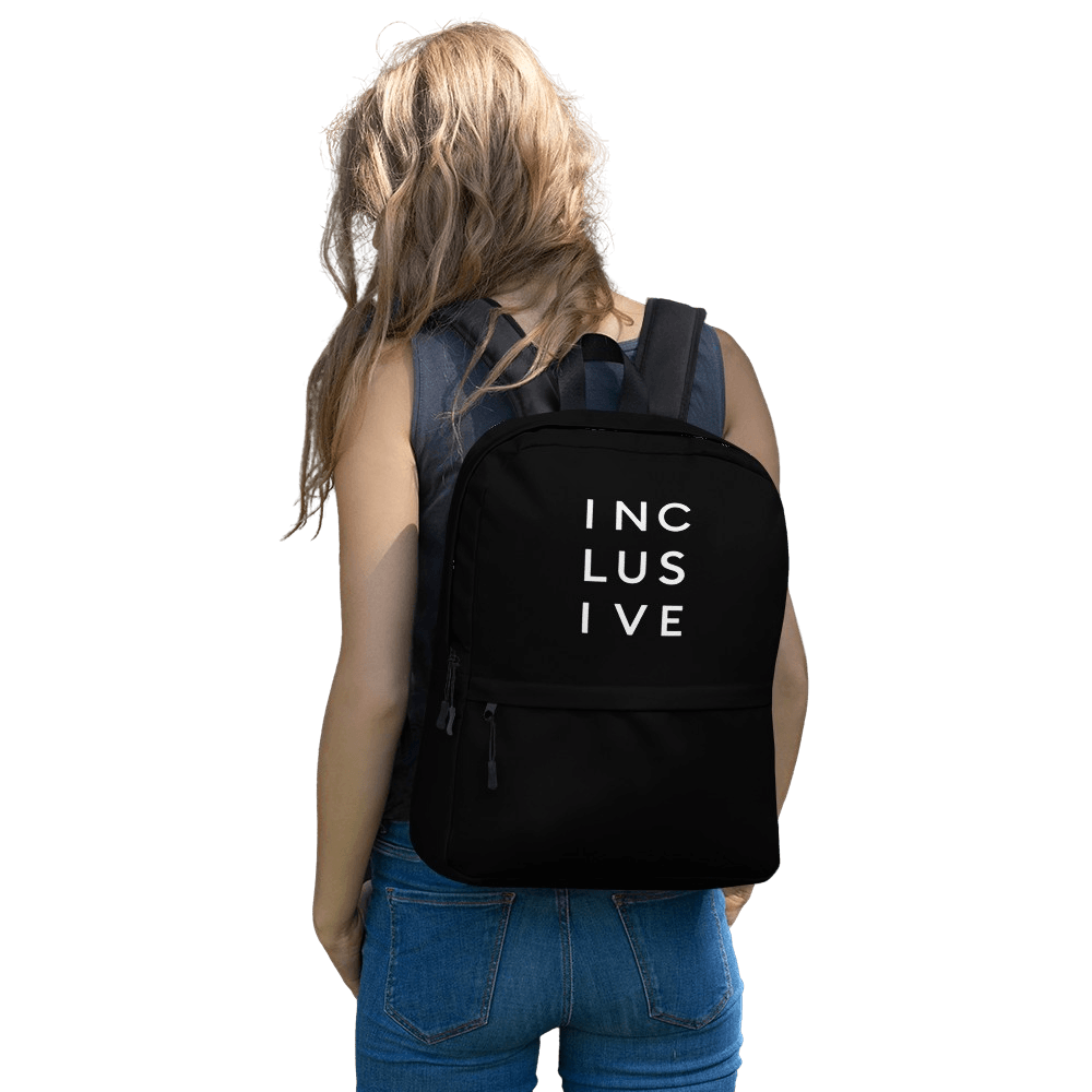 This is a photo of URevolution's black Inclusive backpack. In the top half of the backpack, the word inclusive is printed in white upper case letters evenly spaced over three lines: INC LUS IVE. The rest of the backpack is black. The backpack is being worn on the back of model with long fawn hair, wearing a navy tank top and blue jeans.