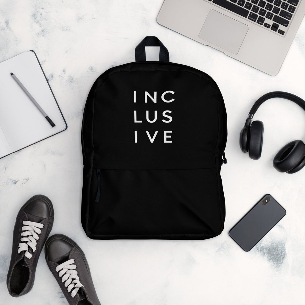 This is a photo of URevolution's black Inclusive backpack. In the top half of the backpack, the word inclusive is printed in white upper case letters evenly spaced over three lines: INC LUS IVE. The rest of the backpack is black. The backpack is in the middle of table. Around the backup is a laptop, headphones, iPhone, and a notepad.